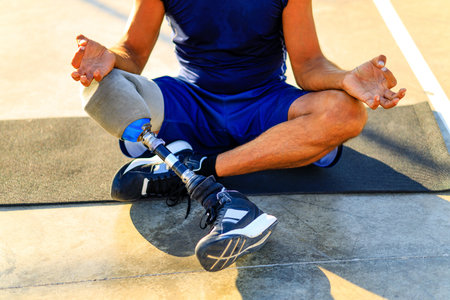 man with prosthesis leg practicing yoga sitting in lotus pose on yoga mat meditating outdoors in sun...