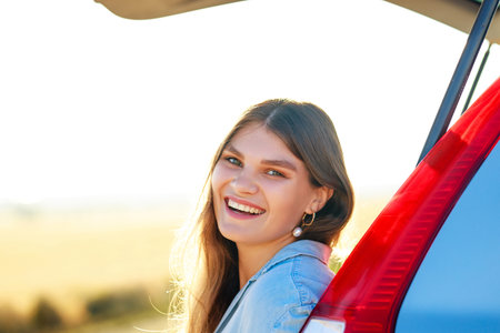 Happy young woman looking at camera near car at golden sunset in fieldの写真素材