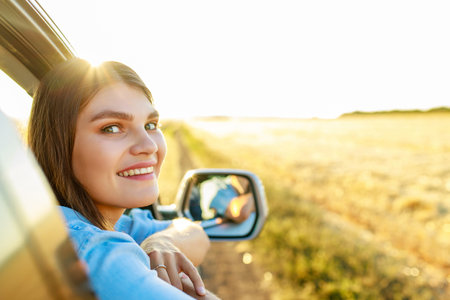 Happy young woman looking out of car window at golden sunset in fieldの写真素材