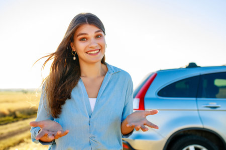 young woman stands with a guilty expression in front of a car in a fieldの写真素材