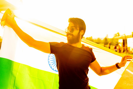 handsome indian bearded man in black cotton t-shirt wawing indian flag in summer park at sunset golg...
