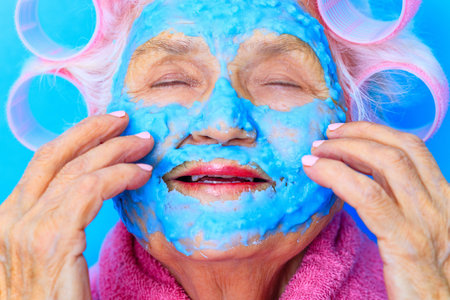 old woman wirh pink curlers on head and blue hyaluronic beauty mask on face wear cotton robe, preparing for a date Valentines Day in studio baclgroundの写真素材