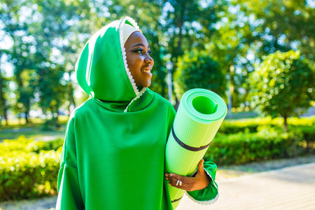 latin black woman in green hijab with bright make up and piercing nose holg yoga mat outdoor in summer parkの写真素材