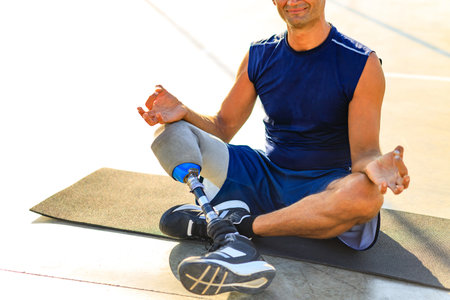 man with prosthesis leg practicing yoga sitting in lotus pose on yoga mat meditating outdoors in sun lightsの写真素材