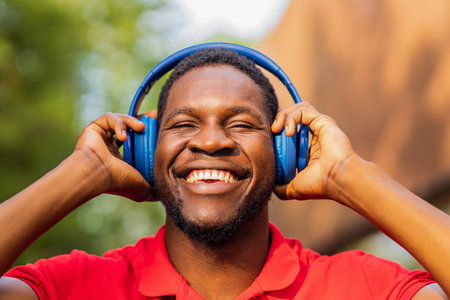 african american man in red t-shirt listening music in blue headphones and being in high spirit outdoorsの写真素材