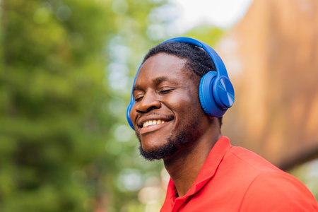 african american man in red t-shirt listening music in blue headphones and being in high spirit outdoorsの写真素材