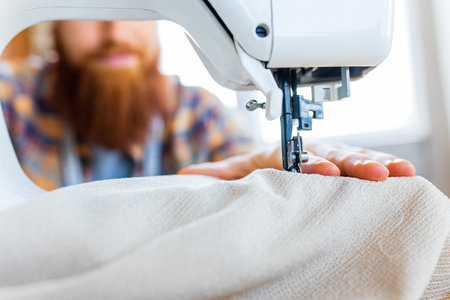 handsome redhaired man with long beard sews at a sewing machine at home studioの写真素材