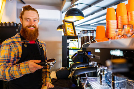 Professional barista young redhaired ginger bearded man in black apron working in coffee shopの写真素材