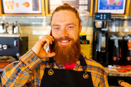 Professional barista young redhaired ginger bearded man in black apron working in coffee shopの写真素材