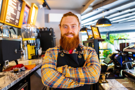Professional barista young redhaired ginger bearded man in black apron working in coffee shopの写真素材
