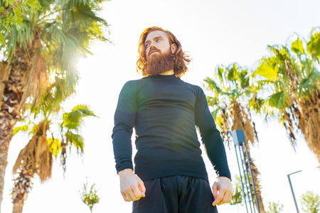 handsome redhaireb man with long beard wear black sporty suit and warming up in sunny beachの写真素材