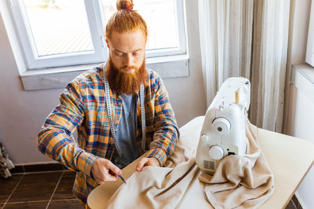 handsome redhaired man with long beard sews at a sewing machine at home studioの写真素材