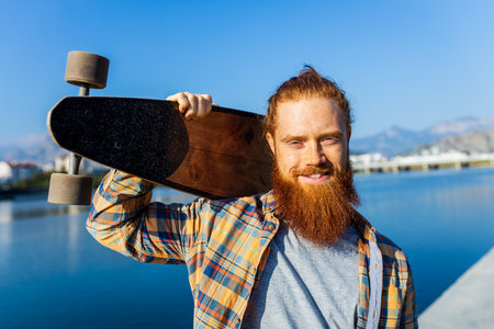 handsome redhaired man with long beard skateboarding near river at sunny summer dayの写真素材