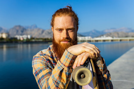 handsome redhaired man with long beard skateboarding near river at sunny summer dayの写真素材