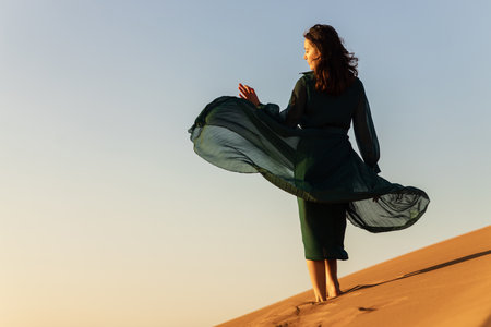 woman in green long dress stands in desert sand dunes background yellow orange sunsetの写真素材