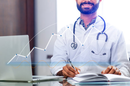A man in a white lab coat is sitting at a desk with a laptop and a notebookの写真素材
