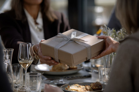 A female executive presents a beautifully wrapped gift box during a business gathering, surrounded by colleagues enjoying a sophisticated dining experience.の素材
