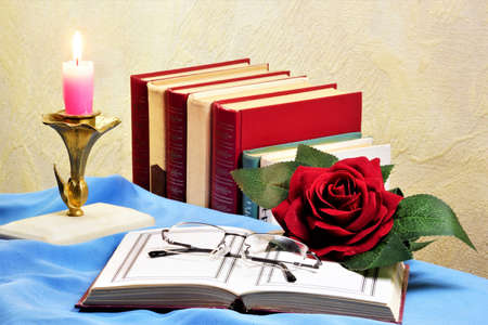 The book is open against the background of a vintage candle, reading glasses and a rose flower...
