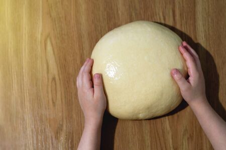 The child holds with his hands a fresh ball of dough on a wooden table. View from above.