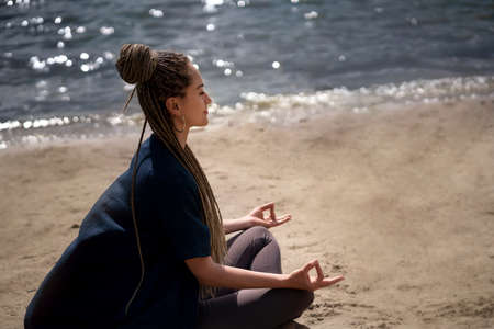 A young girl with an afro braid in sportswear in a lotus position meditates on the beach. River and beautiful cloudy sky. Health and yoga concept.の写真素材
