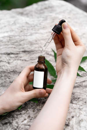 Hands of a young woman with a bottle of serum and a pipette on a marble background. Natural...