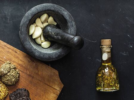 Top view of black mortar and pestle with fresh pieces of garlic inside. Bottle of olive oil with spices. Wooden cutting board with three types of sauces, including french mustard on black chalkboard.の写真素材