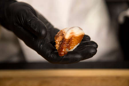 Chef hold in hand traditional sushi nigiri with eel, rice and nori. Close up of chefâs hand in black gloves near wooden board. Japanese food in restaurant. Cooking process of Asian dish preparationの写真素材