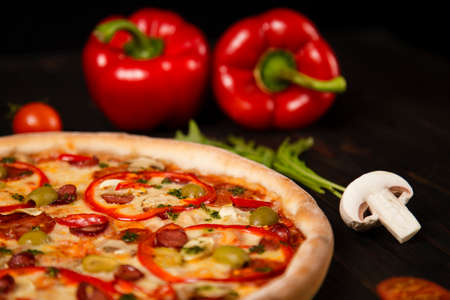 Side view of pizza with bell pepper, green olives, smoked sausages, mozzarella cheese, pepperoni, salami, chili pepper, champignons on wooden board with ingredients on background. uncut tasty pizzaの写真素材