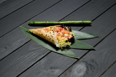 Japanese Sushi Temaki Hand Roll with salmon, rice, Tobiko roe eggs, avocado and salad mix in Mamenori seaweed. Close up of Pan Asian traditional dish on bamboo leaves near bamboo stick on wooden boardの写真素材