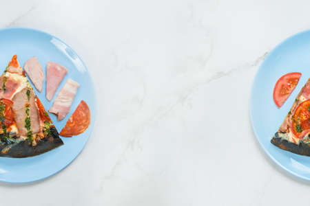 Slices of meat pizza with pesto sauce tomato, salami, ham, bacon, beef and pork fillet on black dough. Pizza with ingredients on pastel blue plates, white marble background, parts of serving plates.の写真素材