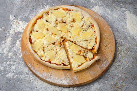 Whole classic Hawaiian Pizza with chicken, pineapple (ananas), mozzarella cheese and tomato sauce served on wooden plate served with culinary flour and spices on gray concrete background.の写真素材