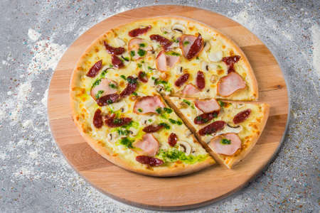Whole rustic Pizza with smoked hunting sausage, ham, mushrooms, mozzarella cheese served with pesto sauce on wooden plate served with culinary flour and spices on gray concrete background.の写真素材