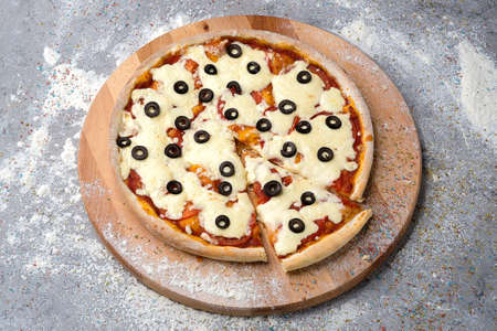 Whole rustic Italian Pizza with tomatoes, black olives (rings) and a lot of mozzarella cheese served on wooden plate served with culinary flour and spices on gray concrete background.の写真素材