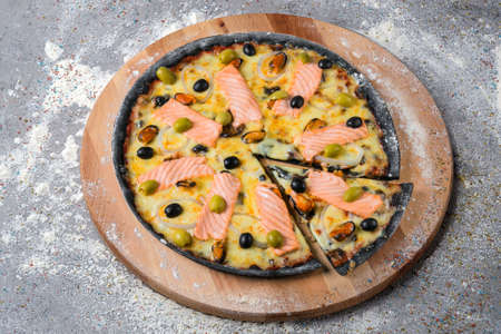 Whole seafood black dough Pizza on wooden plate served with culinary flour and spices on gray concrete background. Fast food Dish With salmon slices, green and black olives and musselsの写真素材