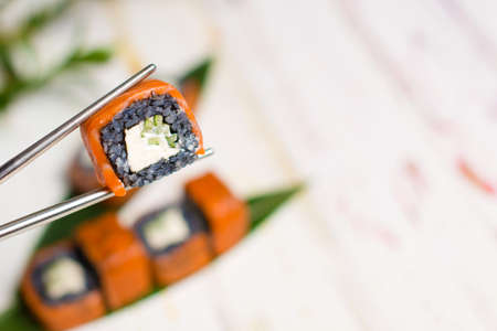 Close up of Sushi Roll piece with black rice and salmon on top in metal chopsticks with blurred background. Philadelphia Sushi Roll with bamboo leaves and plant on background. Copy space.の写真素材