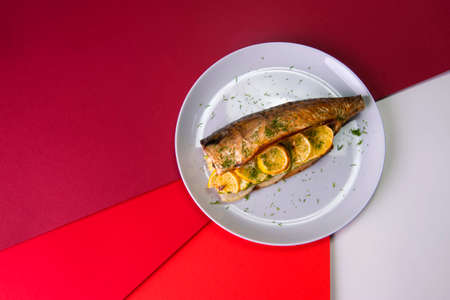 Top view of baked mackerel with lemon slices and onion on gray plate on colorful red background. Grilled fish served with fresh herb dill.の写真素材