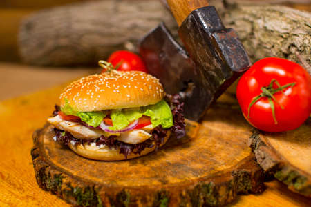 Chicken burger with vegetables and cheese in wheat bun on natural wooden board. Meat Burger with fresh salad leaves, tomatoes, Spanish onions with old rustic axe and wooden logs on backgroundの写真素材