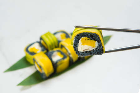 Close up of Sushi Roll piece wrapped in black rice with unagi eel, cream cheese and Tamagoyaki egg omelet inside. hold in metal chopsticks,  blurred Japanese dish with mango and avocado slices on topの写真素材