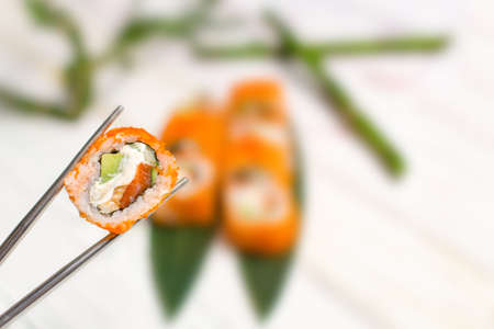 Close up of Sushi Roll piece wrapped in rice and flying fish roe in metal chopsticks with blurred background. Sushi Roll with salmon, eel, avocado. bamboo leaves and plant on background. Copy spaceの写真素材
