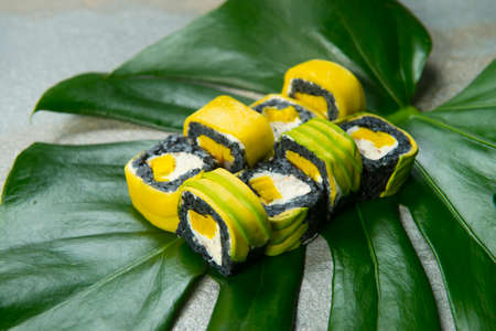Japanese black rice sushi roll with mango, avocado and Philadelphia cheese served on green tropical monstera leaf on anthracite background. Pan Asian menu dish Pieces in breading on exotic plant leafの写真素材