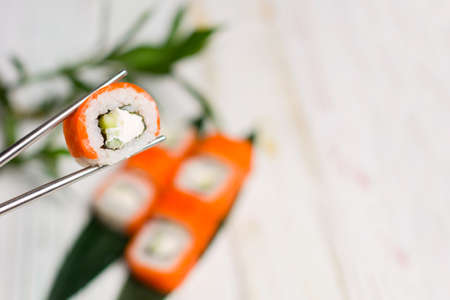 Close up of Sushi Roll piece with salmon on top in metal chopsticks with blurred background. Philadelphia Sushi Roll with bamboo leaves and plant on background. Copy space.の写真素材