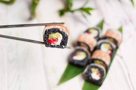 Close up of Sushi Roll piece with black rice and shrimp on top in metal chopsticks with blurred background. Sushi Roll with avocado, cream cheese. bamboo leaves and plant on background. Copy spaceの写真素材
