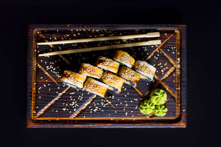 Top view of Japanese sushi roll with black rice, unagi eel served with wasabi sauce, chopsticks and sesame seeds on wooden board. Pan Asian dish on dark backgroundの写真素材