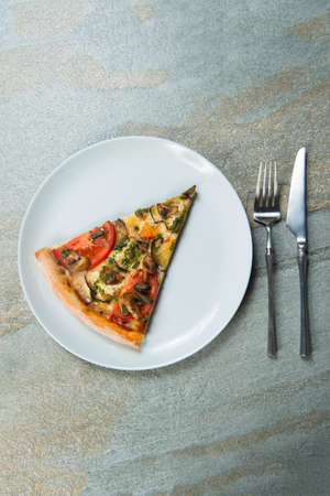 Slice of veggie pizza on plate. Knife and fork near the dish on stone anthracite background. Piece...