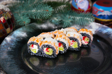 Japanese sushi roll on Christmas decorated plate. Cream cheese, cucumber, red flying fish roe Tobiko wrapped in black rice with black tiger shrimp on top. New year tree brunch and toys on backgroundの写真素材