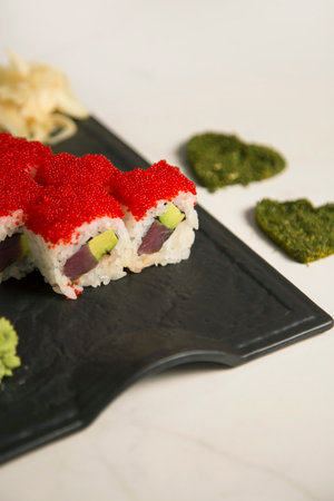 Close up of Japanese sushi roll pieces with tuna, avocado wrapped in rice with red flying fish roe Tobiko on top Black plate served with wasabi, ginger. hearts of dried herbs spices, marble background