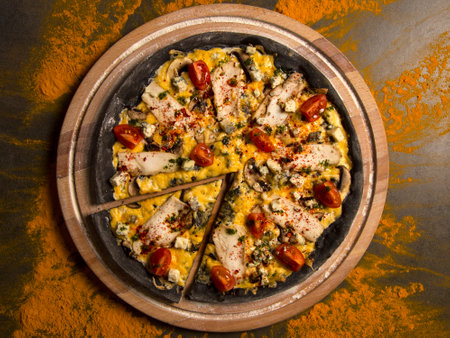 Directly above of black dough pizza served on wooden board. Slice of meat pizza with chicken, tomatoes, blue cheese, champions and melted Gouda cheese. Black chalkboard, scattered turmeric spicesの写真素材
