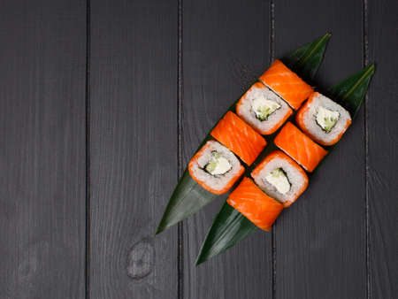 Top view of Japanese sushi roll with raw salmon fillet on top served on bamboo leaves, black wooden background. Cucumber and Philadelphia cream cheese wrapped in rice. Single object, copy text spaceの写真素材