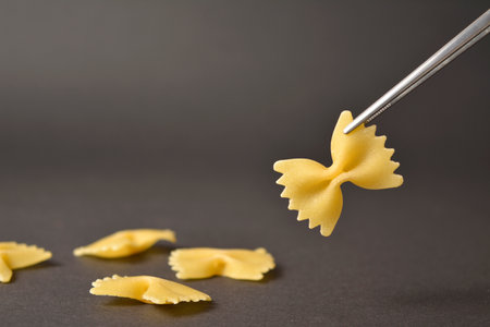 Piece of Uncooked italian farfale butterfly pasta in metal tweezers isolated on dark background with four pieces of raw pasta on backgroundの写真素材