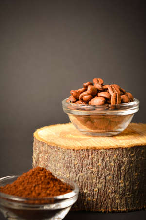 Close-up to Roasted coffee beans served in glass bowl on wooden log, dark background. High quality organic aroma seeds. Ground coffee in bowl out of focus. Copy space text imageの写真素材